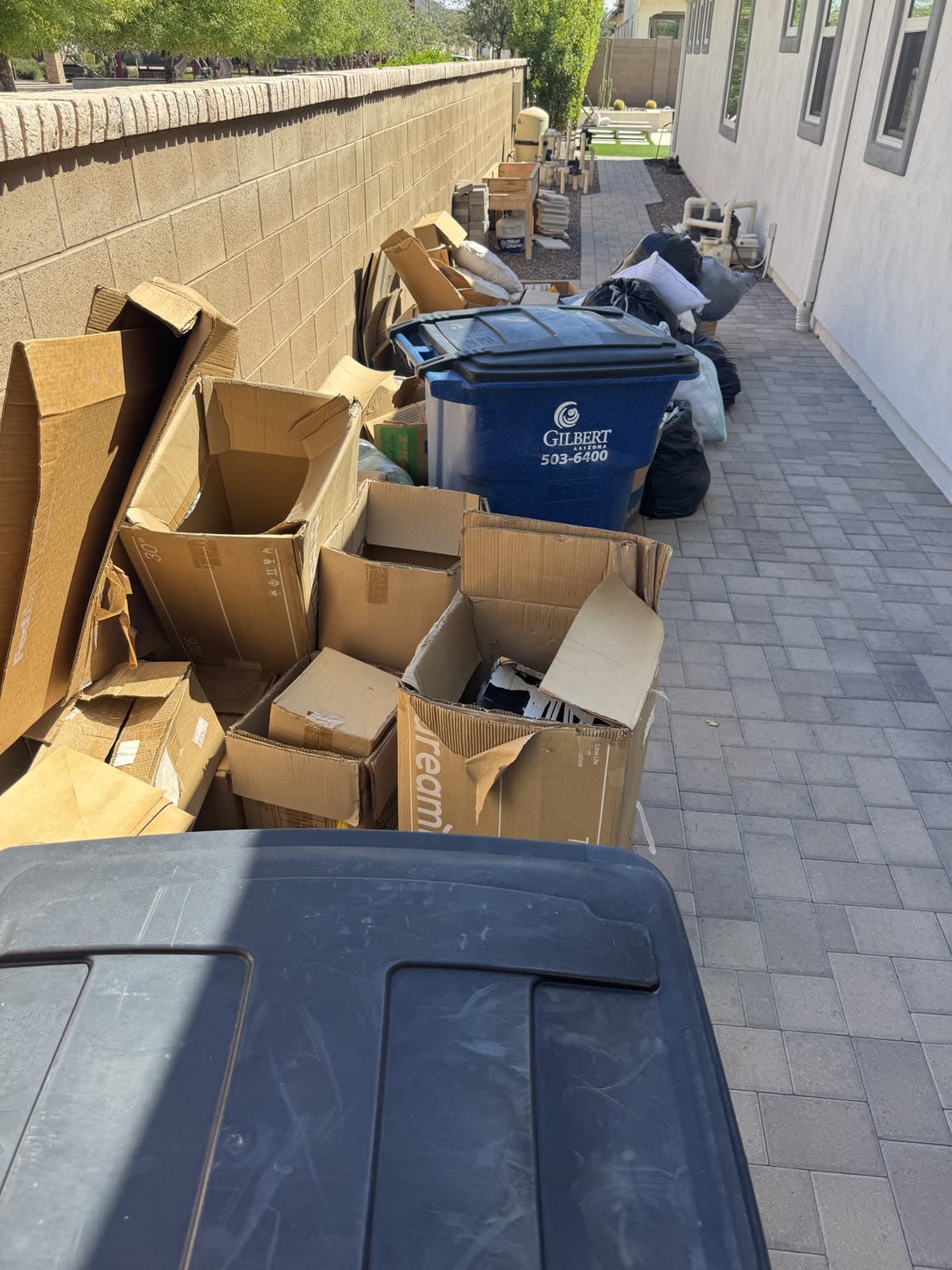 Cardboard boxes and bags stacked down a side yard next to trash bins in Gilbert