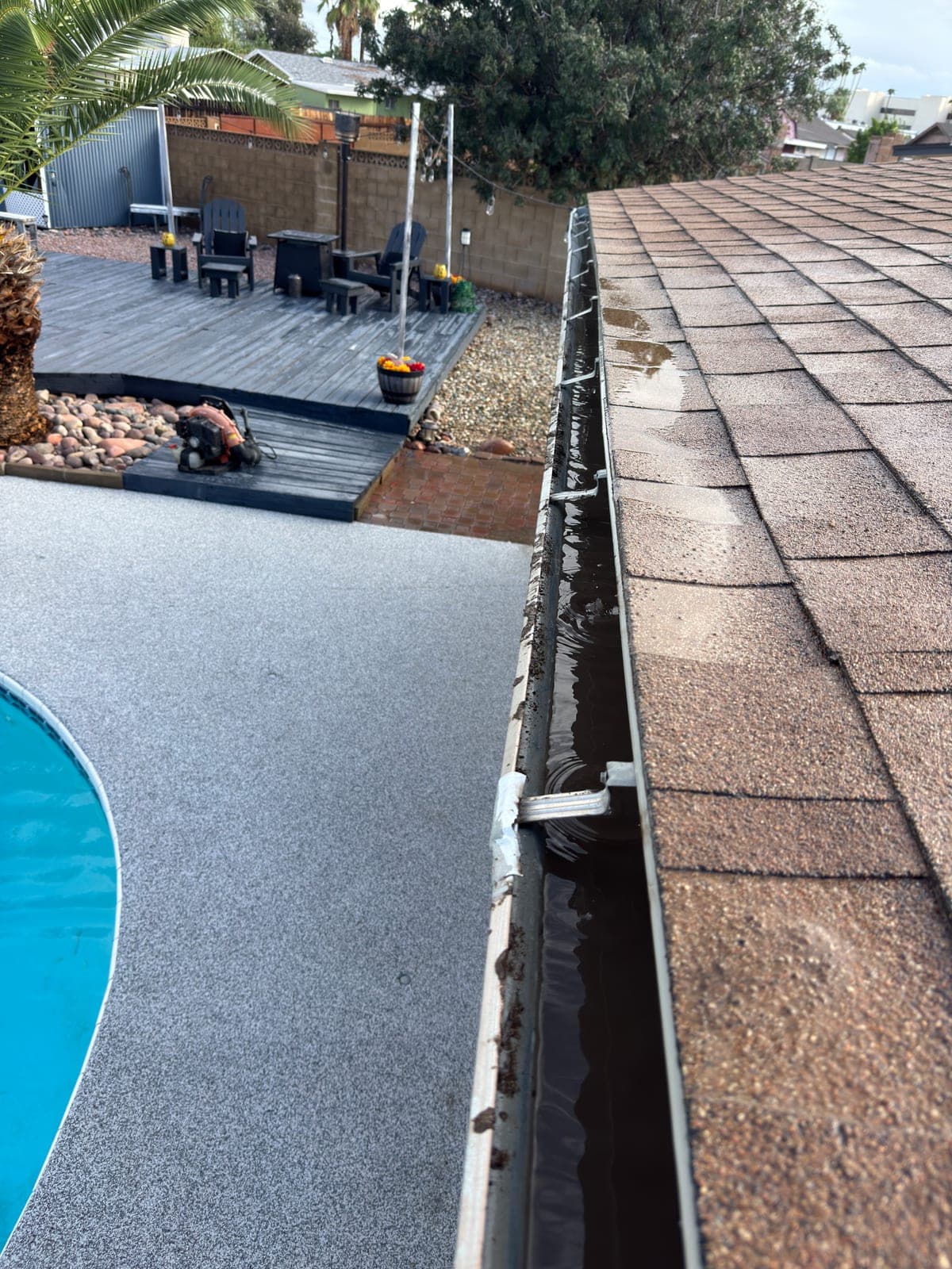 Rain gutter along a shingle roof holding standing water and silt, with a pool and patio deck below